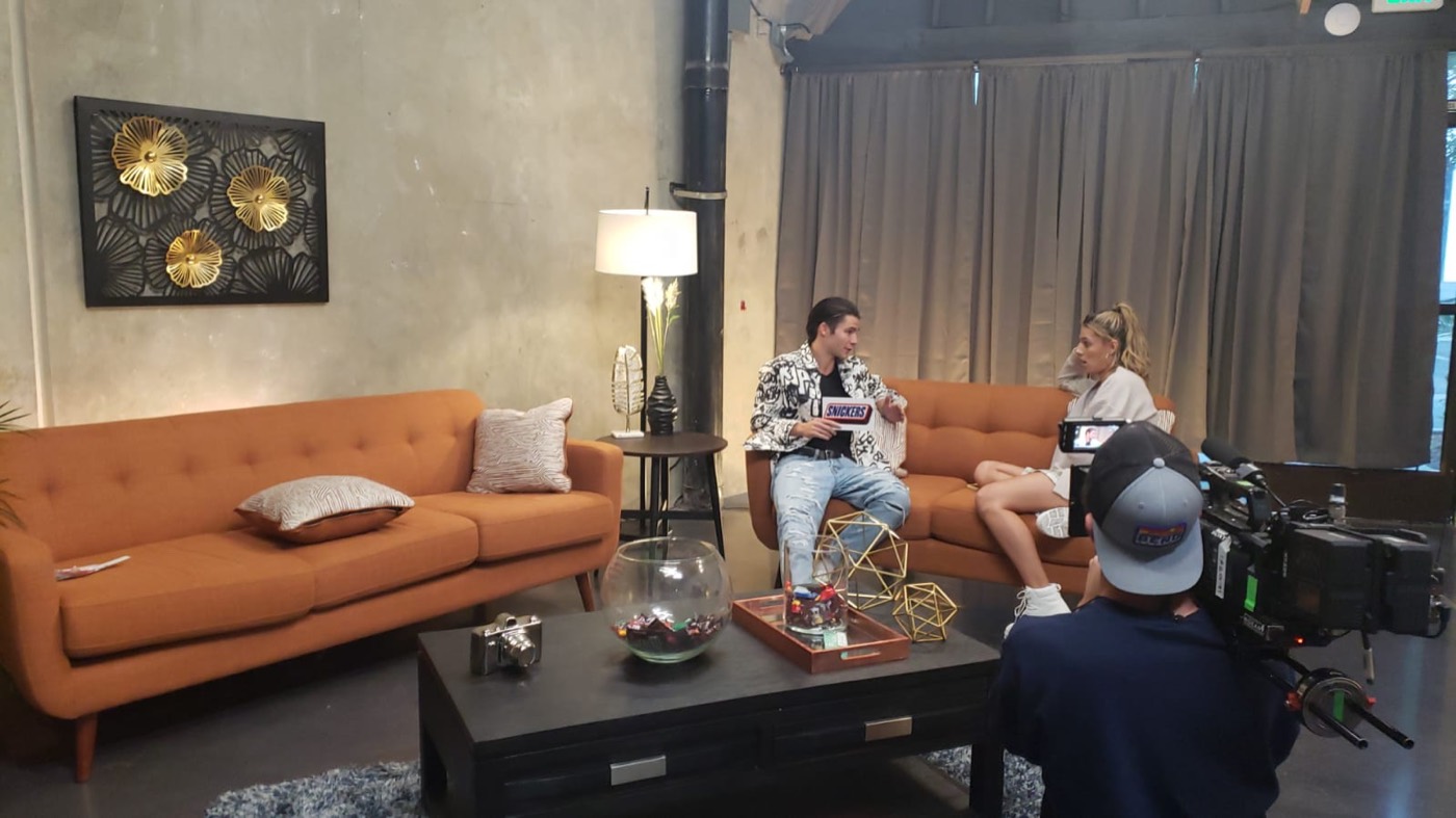 Two hosts on an orange sofa set with a camera operator shooting handheld