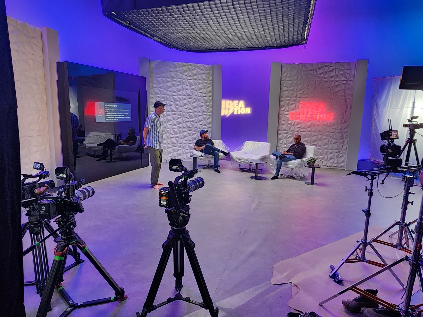 A multi-camera talk-show set under purple light with neon signage